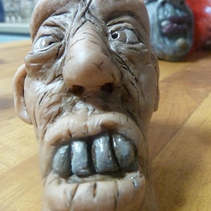 May include: A detailed sculpture of a grotesque face with exaggerated features. The face is a light brown color with deep wrinkles, a large nose, and prominent, discolored teeth. Another similar sculpture is visible in the background.
