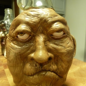 May include: A unique, handcrafted bottle with a sculpted face design. The bottle features a detailed, aged face with a glass top and handle. The face is painted in shades of brown and gold, with expressive eyes and a textured surface.