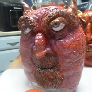 May include: A clear glass bottle decorated with a sculpted, textured red face featuring two small horns, a brown beard, and light blue eyes. The sculpture covers the front of the bottle, which has a glossy finish and a detailed, wrinkled skin texture.