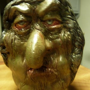 May include: A grotesque, aged face sculpture with a large nose, red-rimmed eyes, and a grimacing mouth. The sculpture is primarily olive green with metallic silver accents, suggesting an antique or fantasy theme. The face is part of a jug.