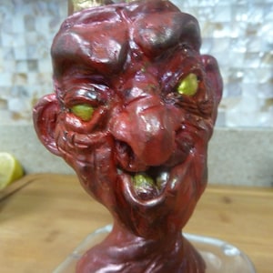 May include: A red devil figurine with golden horns and yellow eyes. The sculpture has a detailed, textured surface and a wide, toothy grin. The figurine is mounted on a clear glass base.