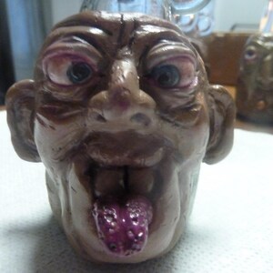 May include: A brown ceramic mug shaped like a grotesque human head with large ears, a prominent nose, and deep-set eyes. The figure has a wide mouth with a textured, purple tongue protruding outward. The surface has a glossy finish with visible details.