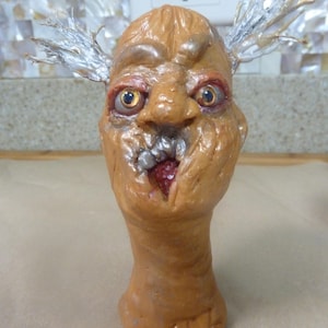 May include: A grotesque, tan-colored sculpture of a face with exaggerated features. The face has wide, staring eyes, a large nose, and an open mouth revealing teeth. Silver-colored, spiky protrusions extend from the head. The sculpture is mounted on a neck-like base.