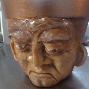 May include: A ceramic planter shaped like a man's head with a distressed, aged look. The planter has a copper-colored rim and a textured surface.