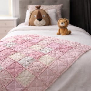 May include: A handmade rag quilt featuring a patchwork design of square fabric blocks in various shades of pink, white, and floral patterns. The quilt has exposed, frayed seams between each square and a quilted X pattern on every individual block.
