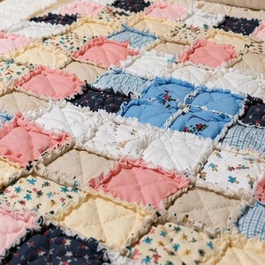 May include: A patchwork quilt in pastel shades. The quilt is made up of squares of pink, blue, white, and yellow fabrics, some with floral patterns. The edges of the squares are ruffled, adding texture. The quilt is a cozy, handmade item.