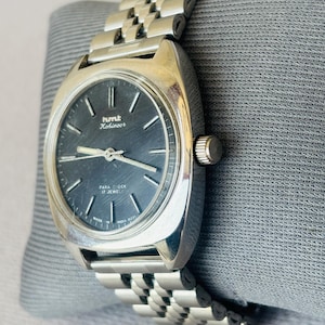Puede incluir: Un reloj de pulsera vintage plateado con una esfera gris oscuro y una correa de metal a juego. La esfera del reloj muestra la marca "HMT Kohinoor" y el texto "Para Shock 17 Jewels". El reloj est&aacute; sobre una superficie gris.