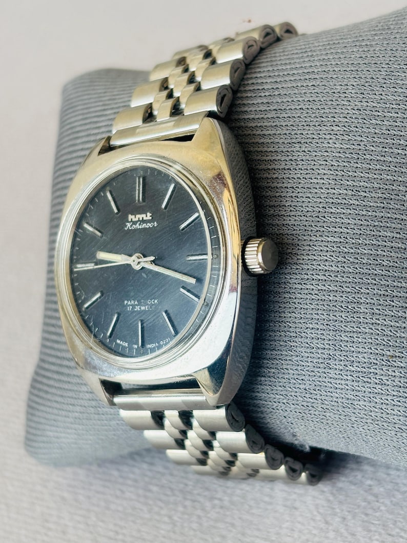 Puede incluir: Un reloj de pulsera vintage plateado con una esfera gris oscuro y una correa de metal a juego. La esfera del reloj muestra la marca "HMT Kohinoor" y el texto "Para Shock 17 Jewels". El reloj est&aacute; sobre una superficie gris.