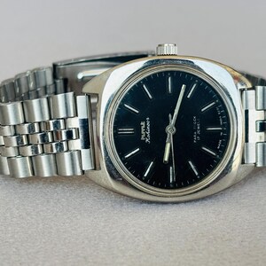 Puede incluir: Un reloj de pulsera plateado con esfera negra y correa de eslabones plateados. La esfera del reloj tiene marcadores de hora y agujas plateadas. El reloj est&aacute; etiquetado como "HMT Kohinoor" y "17 Jewels".