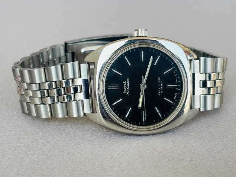 Puede incluir: Un reloj de pulsera plateado con esfera negra y correa de eslabones plateados. La esfera del reloj tiene marcadores de hora y agujas plateadas. El reloj est&aacute; etiquetado como "HMT Kohinoor" y "17 Jewels".