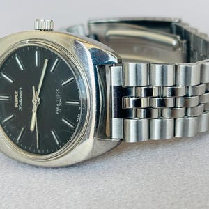 Puede incluir: Un reloj de pulsera plateado vintage con esfera negra. El reloj tiene una correa plateada y una esfera redonda con marcadores y agujas blancas. La esfera del reloj muestra el texto "Kmt Kohinoor" y "Para Shock 17 Jewels".