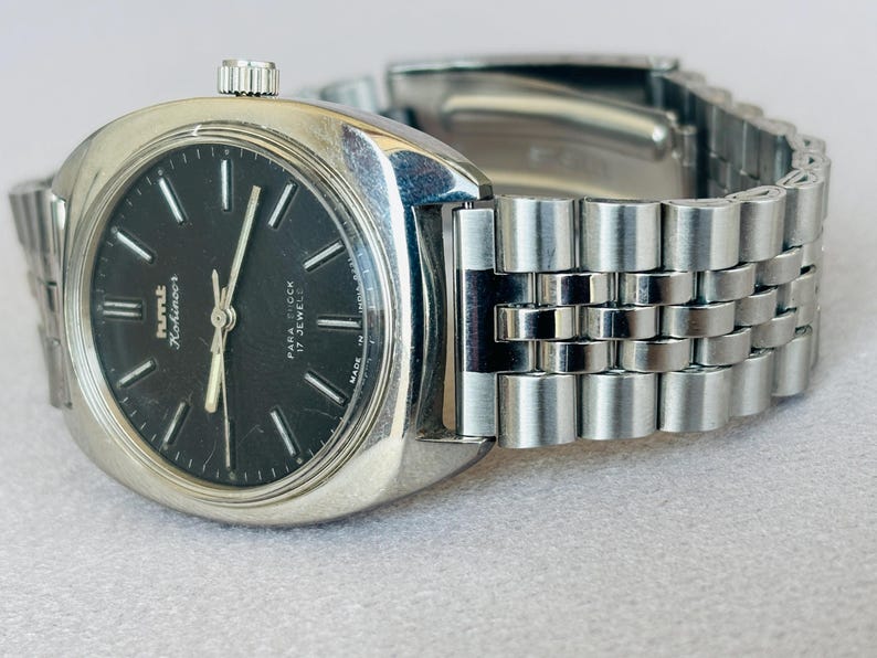 Puede incluir: Un reloj de pulsera plateado vintage con esfera negra. El reloj tiene una correa plateada y una esfera redonda con marcadores y agujas blancas. La esfera del reloj muestra el texto "Kmt Kohinoor" y "Para Shock 17 Jewels".