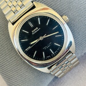 Puede incluir: Un reloj de pulsera HMT Kohinoor vintage con caja y correa plateadas. El reloj tiene una esfera negra con marcadores y agujas plateadas. La esfera indica "PARA SHOCK 17 JEWELS" y "MADE IN INDIA".