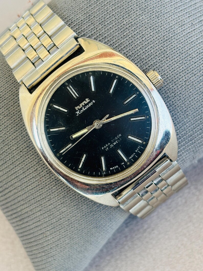 Puede incluir: Un reloj de pulsera HMT Kohinoor vintage con caja y correa plateadas. El reloj tiene una esfera negra con marcadores y agujas plateadas. La esfera indica "PARA SHOCK 17 JEWELS" y "MADE IN INDIA".