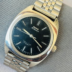 Puede incluir: Un reloj de pulsera vintage con una correa y caja de metal plateado. La esfera del reloj es negra con manecillas e indicadores plateados. El reloj tiene el texto "hmt Kohinoor" y "Para Shock 17 Jewels". Hecho en India.