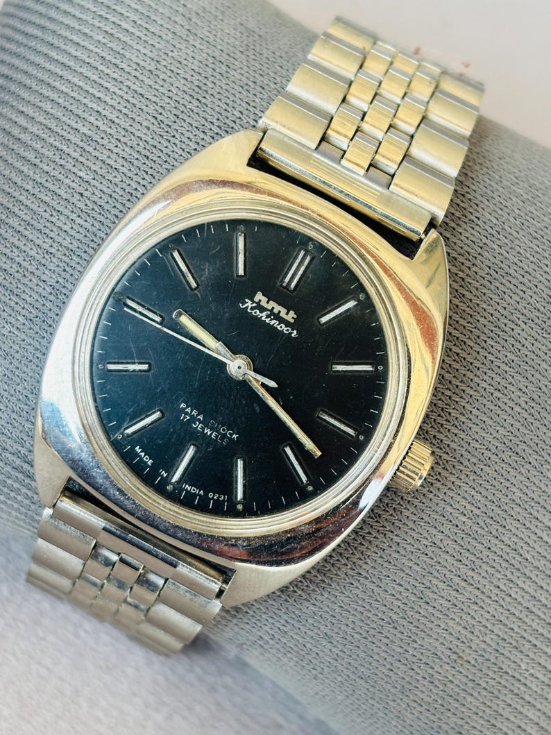 Puede incluir: Un reloj de pulsera vintage con una correa y caja de metal plateado. La esfera del reloj es negra con manecillas e indicadores plateados. El reloj tiene el texto "hmt Kohinoor" y "Para Shock 17 Jewels". Hecho en India.