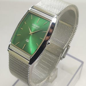 May include: A rectangular Seiko Quartz wristwatch featuring a vibrant green sunray dial with gold-tone hour markers and hands. The watch has a polished silver-tone case and a flexible silver-tone stainless steel mesh strap with a secure clasp.