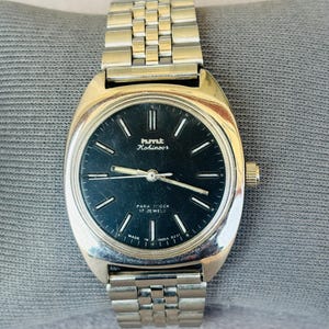 Puede incluir: Un reloj de pulsera plateado con esfera negra. El reloj tiene una caja rectangular y una correa de metal. La esfera muestra el texto "hmt Kohinoor" y "PARA SHOCK 17 JEWELS". El reloj est&aacute; sobre una superficie gris.