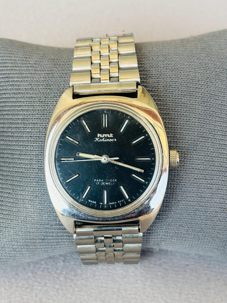 Puede incluir: Un reloj de pulsera plateado con esfera negra. El reloj tiene una caja rectangular y una correa de metal. La esfera muestra el texto "hmt Kohinoor" y "PARA SHOCK 17 JEWELS". El reloj est&aacute; sobre una superficie gris.