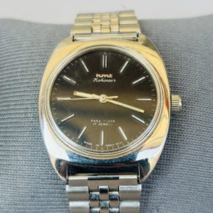 Puede incluir: Reloj de pulsera HMT Kohinoor plateado con esfera oscura y agujas e indicadores plateados. El reloj tiene una correa plateada y una corona en el lado derecho. La esfera incluye el texto "PARA SHOCK 17 JEWELS" y "MADE IN INDIA 0231".