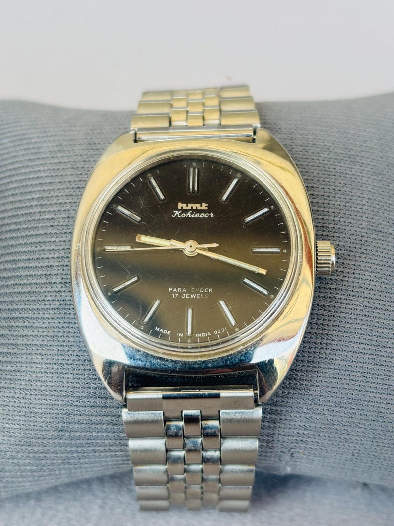 Puede incluir: Reloj de pulsera HMT Kohinoor plateado con esfera oscura y agujas e indicadores plateados. El reloj tiene una correa plateada y una corona en el lado derecho. La esfera incluye el texto "PARA SHOCK 17 JEWELS" y "MADE IN INDIA 0231".
