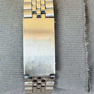 Puede incluir: Primer plano de una correa de reloj plateada con un cierre rectangular. La correa est&aacute; compuesta por peque&ntilde;os eslabones rectangulares. El cierre tiene un acabado de metal cepillado y un dise&ntilde;o sutil grabado. La correa del reloj descansa sobre una tela gris.