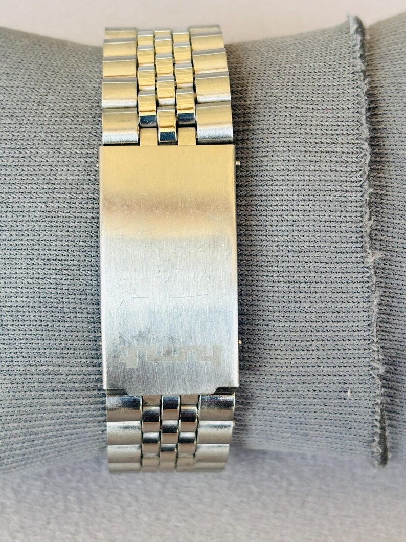 Puede incluir: Primer plano de una correa de reloj plateada con un cierre rectangular. La correa est&aacute; compuesta por peque&ntilde;os eslabones rectangulares. El cierre tiene un acabado de metal cepillado y un dise&ntilde;o sutil grabado. La correa del reloj descansa sobre una tela gris.