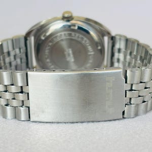 Puede incluir: Primer plano de un reloj de pulsera plateado con una correa de acero inoxidable. El reloj tiene una esfera redonda y un cierre con un dise&ntilde;o texturizado. La correa est&aacute; hecha de peque&ntilde;os eslabones met&aacute;licos conectados.