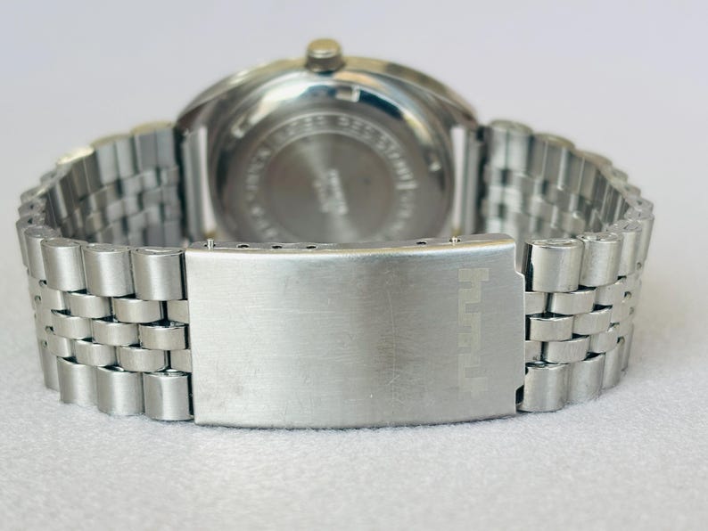 Puede incluir: Primer plano de un reloj de pulsera plateado con una correa de acero inoxidable. El reloj tiene una esfera redonda y un cierre con un dise&ntilde;o texturizado. La correa est&aacute; hecha de peque&ntilde;os eslabones met&aacute;licos conectados.