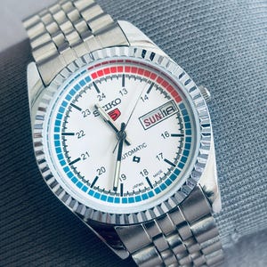 May include: A silver-toned Seiko 5 automatic watch with a white face, featuring red and blue accents. The watch has a stainless steel band and a day-date display. The watch face displays the day as "SUN 18".