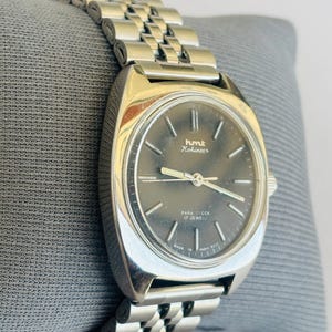 Puede incluir: Un reloj HMT Kohinoor plateado con una esfera gris oscuro y una correa de metal con eslabones. La esfera del reloj tiene agujas e indicadores plateados. El reloj est&aacute; marcado "PARA SHOCK 17 JEWELS" y "MADE IN INDIA 0231".