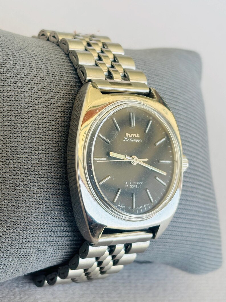 Puede incluir: Un reloj HMT Kohinoor plateado con una esfera gris oscuro y una correa de metal con eslabones. La esfera del reloj tiene agujas e indicadores plateados. El reloj est&aacute; marcado "PARA SHOCK 17 JEWELS" y "MADE IN INDIA 0231".