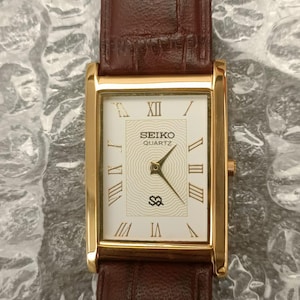 May include: A rectangular gold-tone Seiko Quartz wristwatch with a white dial featuring gold Roman numeral hour markers and a central wavy line pattern. The watch has a brown leather strap with a textured, embossed crocodile-style finish.