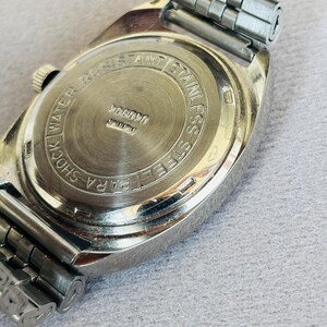 Puede incluir: Primer plano de un reloj vintage de acero inoxidable. La parte trasera del reloj es visible, con el texto "SHOCK WATER RESISTANT STAINLESS STEEL PARA". La correa del reloj es de metal plateado.