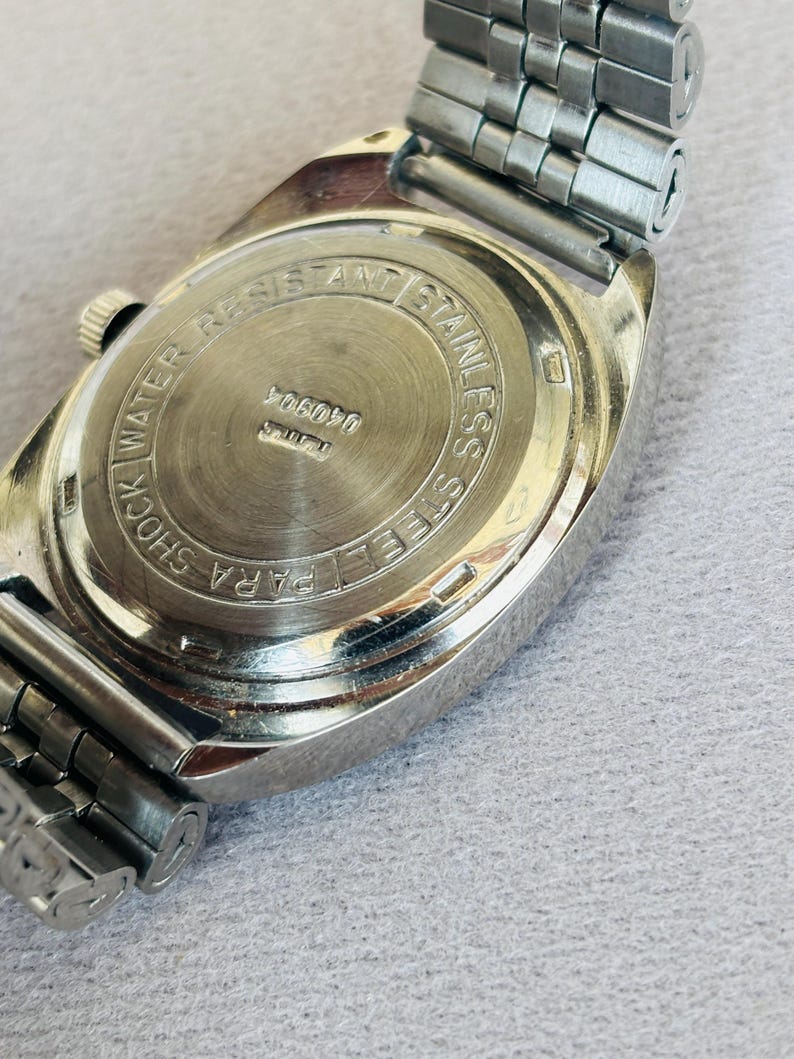 Puede incluir: Primer plano de un reloj vintage de acero inoxidable. La parte trasera del reloj es visible, con el texto "SHOCK WATER RESISTANT STAINLESS STEEL PARA". La correa del reloj es de metal plateado.