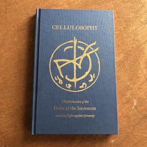 Puede incluir: Un libro de tapa dura azul con el título "CELLULOSOPHY" en letras doradas. La portada presenta un diseño circular dorado con símbolos y el texto "The formation of the Order of the Increscent and our fight against tyranny".