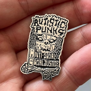 Puede incluir: Un pin plateado con las palabras "AUTISTIC PUNKS" sobre una cara de calavera. Debajo, el pin dice "ANTI-SOCIAL SOCIAL JUSTICE". El pin tiene una estética detallada de punk rock.