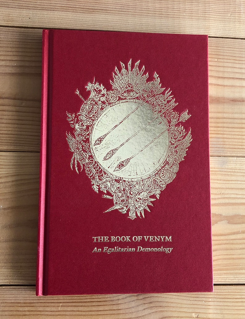 May include: A hardcover book titled "THE BOOK OF VENYM" in red with gold foil accents. The cover features a detailed emblem with arrows and ornate designs. The subtitle reads "An Egalitarian Demonology".