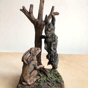 Puede incluir: Una escultura de cerámica de dos conejos, uno marrón y otro gris, con ojos rojos. El conejo gris está colgado de una rama de árbol, mientras que el conejo marrón está sentado en el suelo. La escultura está colocada sobre una base con una superficie texturizada marrón y verde.