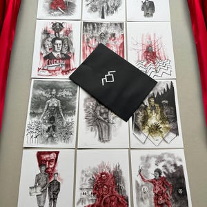 May include: A selection of art prints showcasing surreal and gothic illustrations in monochrome with red highlights. A black envelope with a white geometric design is placed on top. The prints are arranged on a light grey surface.
