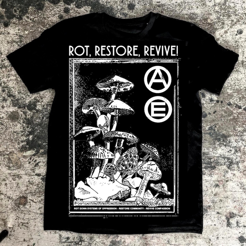 May include: Black t-shirt featuring a white graphic of mushrooms and the text "ROT, RESTORE, REVIVE!" with a circle containing the letter "A" and a circle containing the letter "E". The text below the graphic reads "ROT DOWN SYSTEMS OF OPPRESSION - RESTORE COMMUNITY - REVIVE COMPASSION".