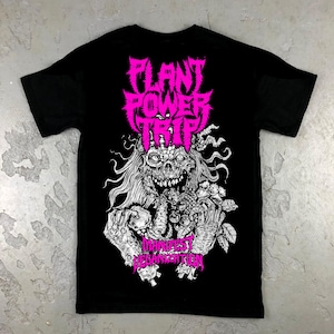 May include: Black t-shirt with a white graphic print of a skull with long hair and vines. The skull is holding a bunch of fruit and vegetables. The text "PLANT POWER TRIP" and "MANIFEST VEGANIZATION" is printed in pink.