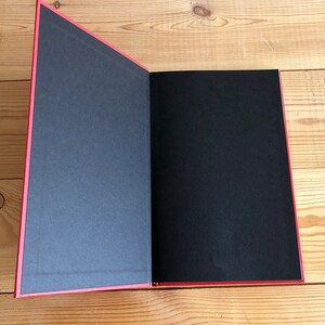 May include: An open book with a red cover and spine, revealing black pages. The pages are blank, and the book is lying on a wooden surface. The edges of the pages are also red.