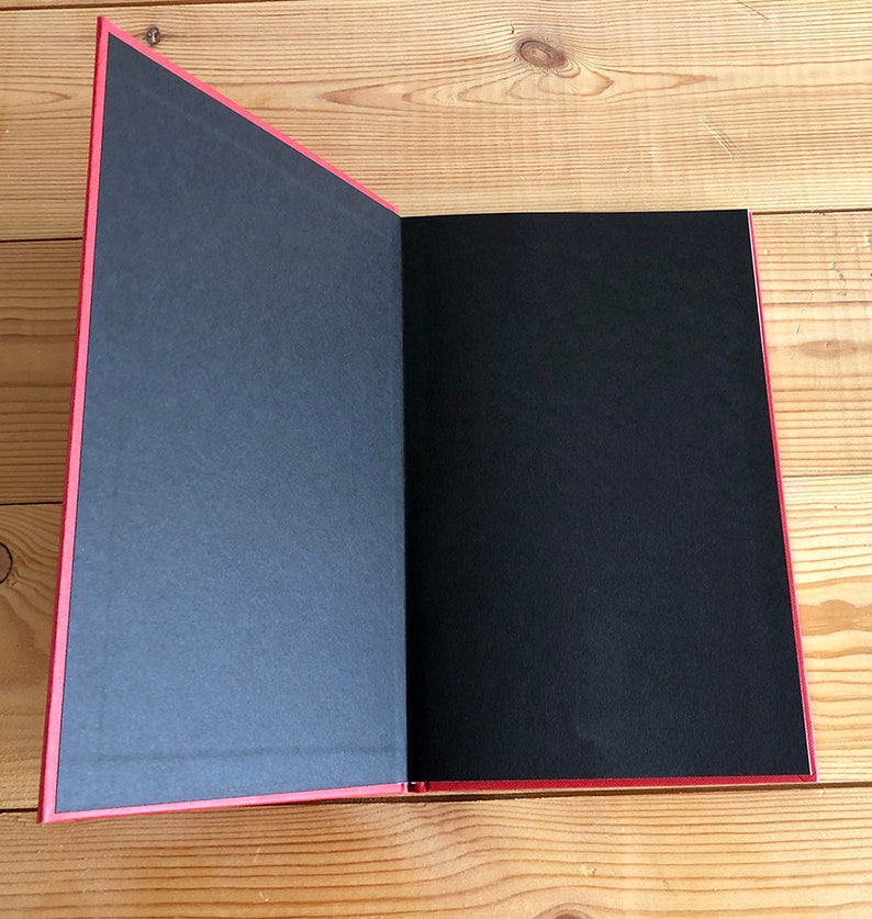 May include: An open book with a red cover and spine, revealing black pages. The pages are blank, and the book is lying on a wooden surface. The edges of the pages are also red.