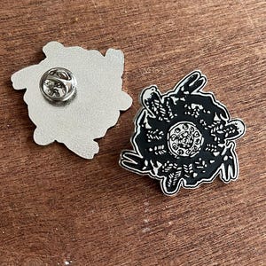 Puede incluir: Un pin de esmalte negro y blanco con un diseño floral detallado. El pin tiene un respaldo plateado con un cierre de alfiler. El pin está sobre una superficie de madera.