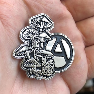 Op de afbeelding: Een zilverkleurige pin met een cluster van gedetailleerde paddenstoelillustraties en een zwart en zilver anarchiesymbool. De pin heeft een gedetailleerd, reliëf ontwerp met een zwarte rand.