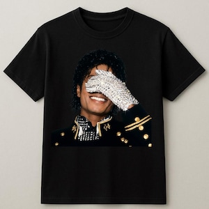 May include: Black t-shirt featuring a portrait of a smiling person with a sequined glove covering one eye. The person is wearing a black jacket with gold accents. The image is a close-up, highlighting the details of the clothing and the person's expression.