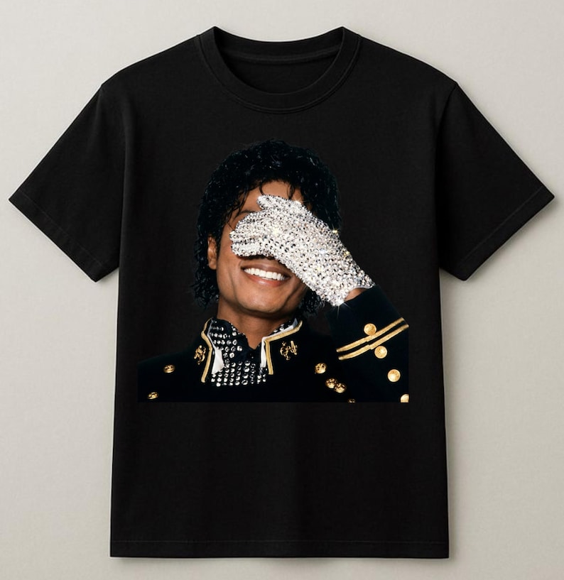 May include: Black t-shirt featuring a portrait of a smiling person with a sequined glove covering one eye. The person is wearing a black jacket with gold accents. The image is a close-up, highlighting the details of the clothing and the person's expression.