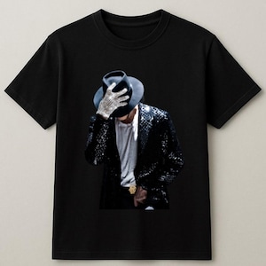 May include: Black t-shirt featuring a graphic of a person wearing a black hat, sequined jacket, and a single jeweled glove. The image is a portrait of a famous musician. The shirt is a classic crew neck style.