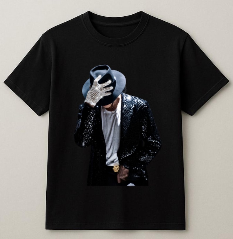 May include: Black t-shirt featuring a graphic of a person wearing a black hat, sequined jacket, and a single jeweled glove. The image is a portrait of a famous musician. The shirt is a classic crew neck style.